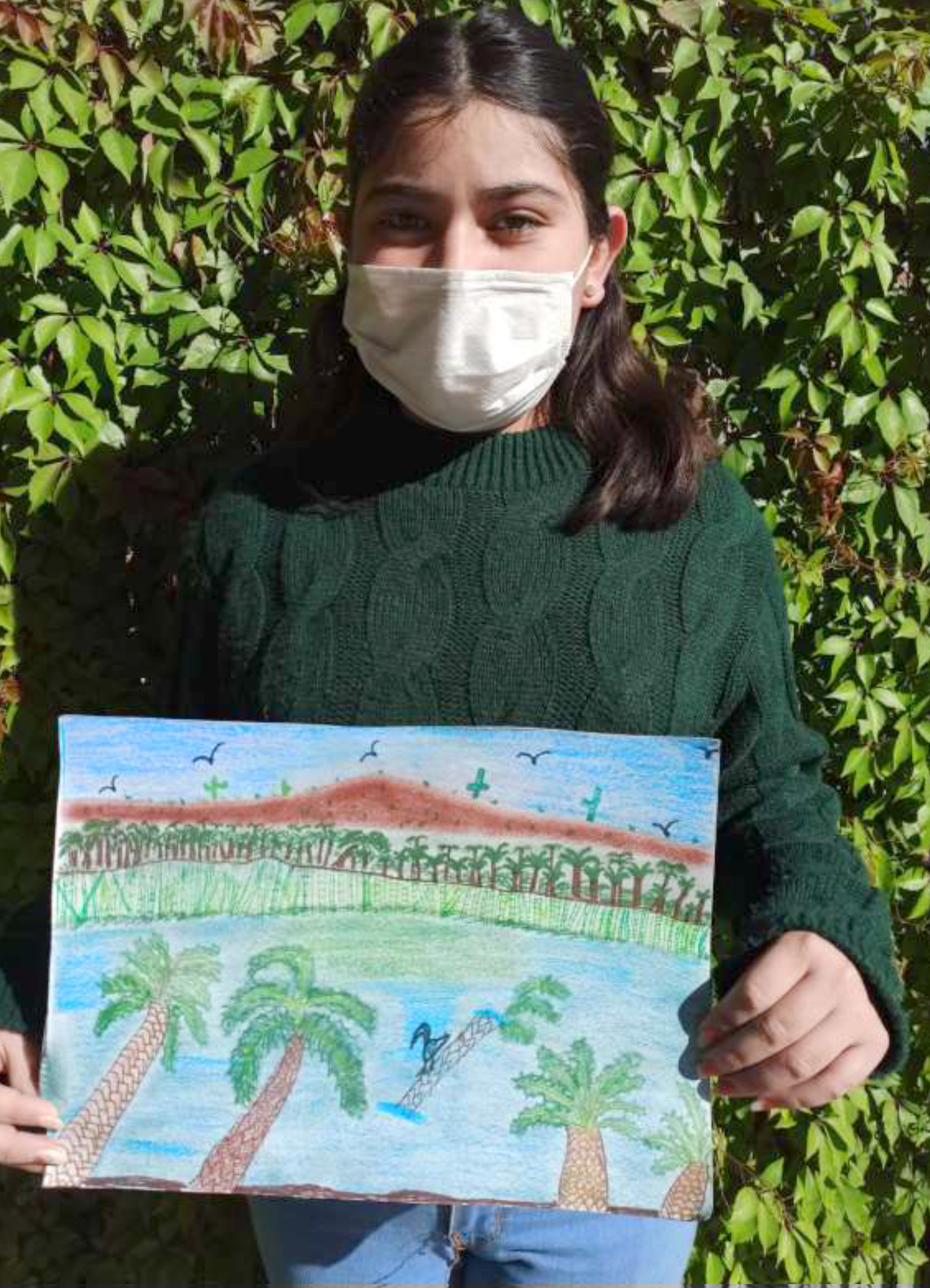 Participante Camila sosteniendo su dibujo con paisaje de palmeras y vegetación rodeando una laguna, con pájaros en el aire y montaña al fondo.